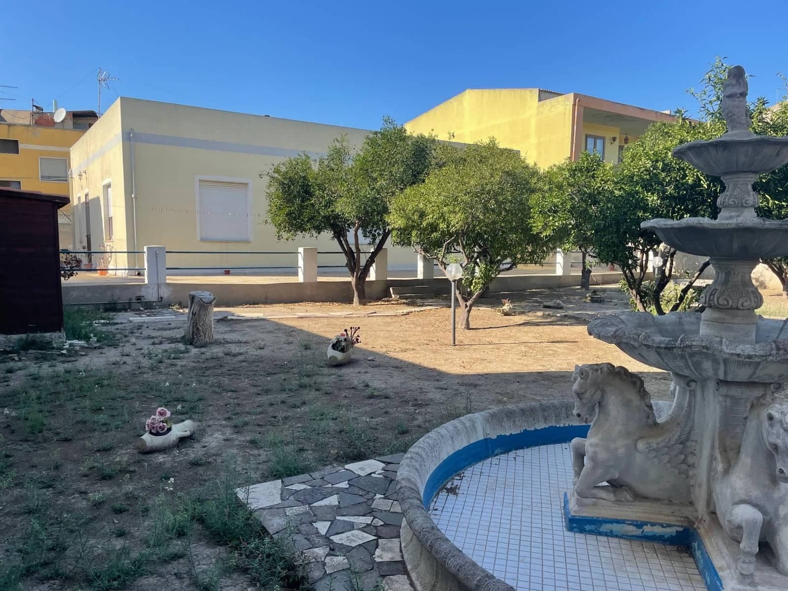 Giardino con alberi e una fontana in pietra a più vasche, davanti a una casa a un piano dalle pareti gialle