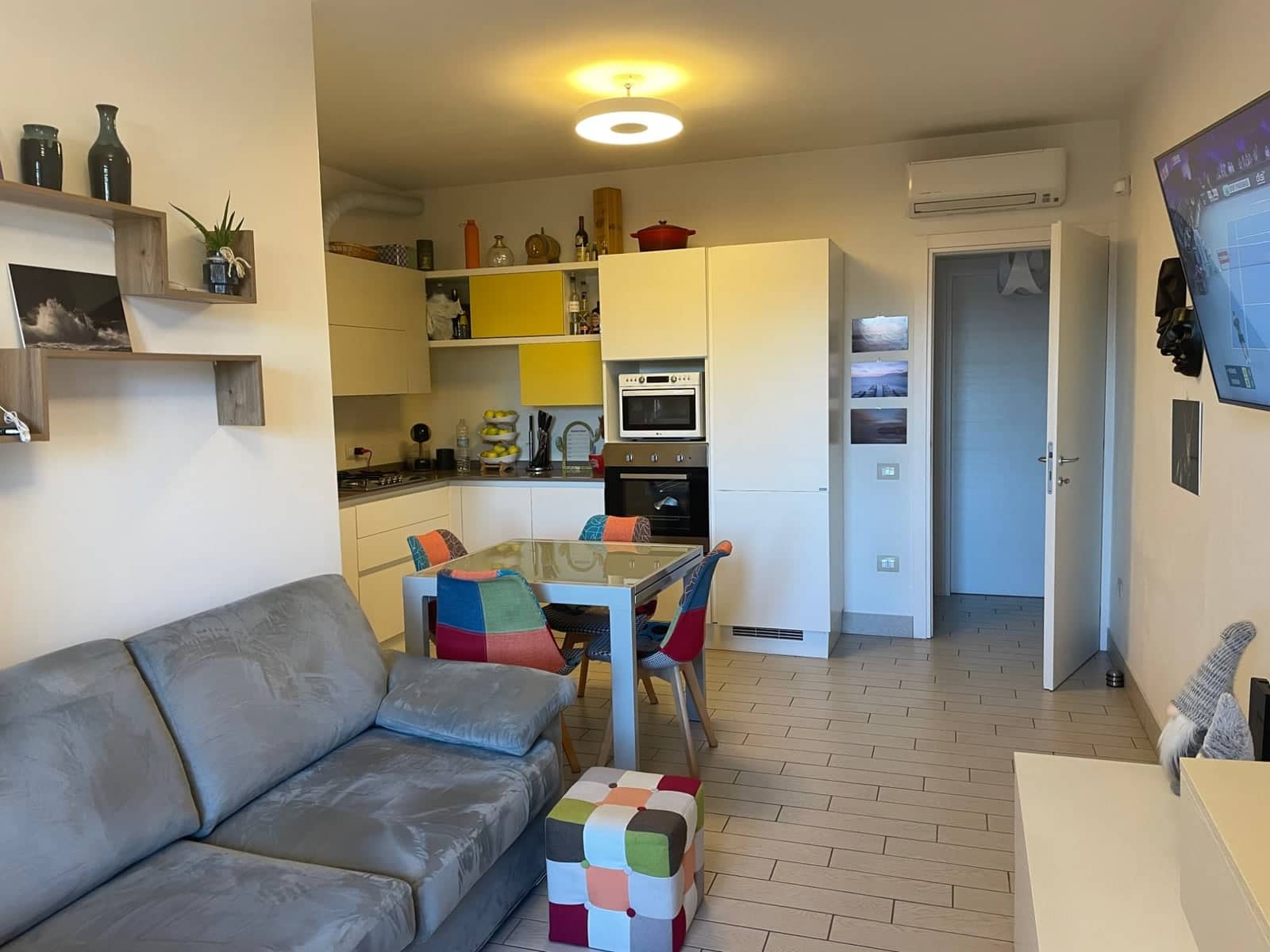 Soggiorno con divano grigio e tavolo da pranzo, aperto sulla cucina con ante bianche e gialle